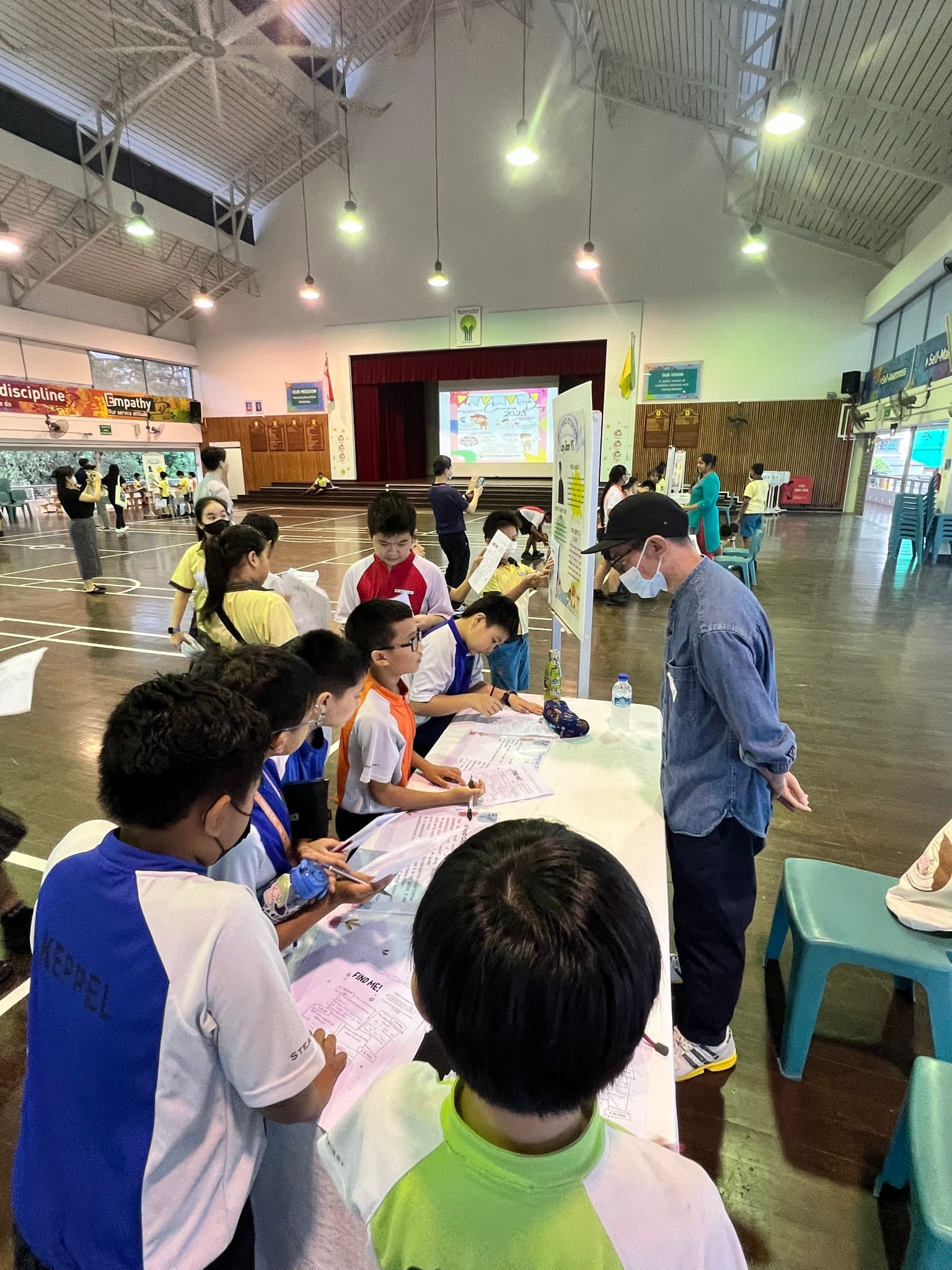 Children gather around a table with worksheets in a school hall, supervised by an adult wearing a mask and cap.